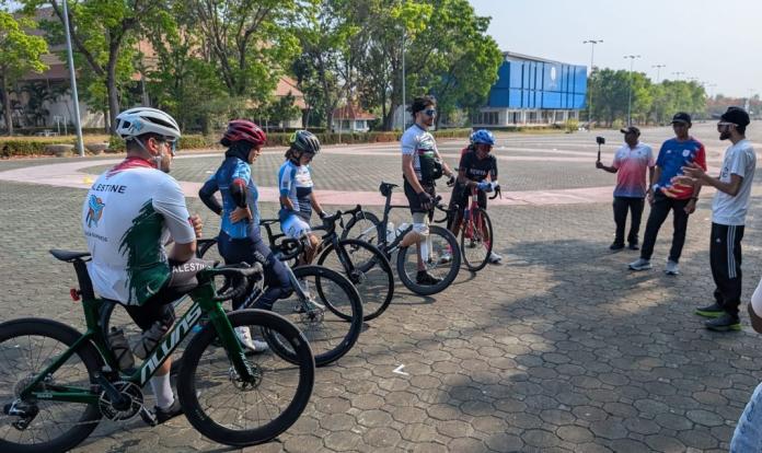 Five people wearing helmets are standing next to their bikes, while listening to an official speaking